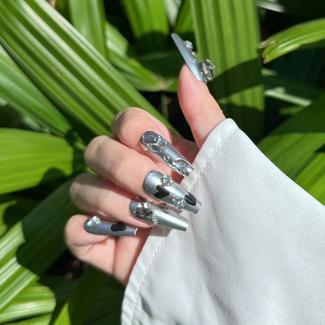 Heart Spell handmade long coffin-shaped metallic silver press-on nails with black heart design, gray heart gems, unique rhinestones, and metal chain accents