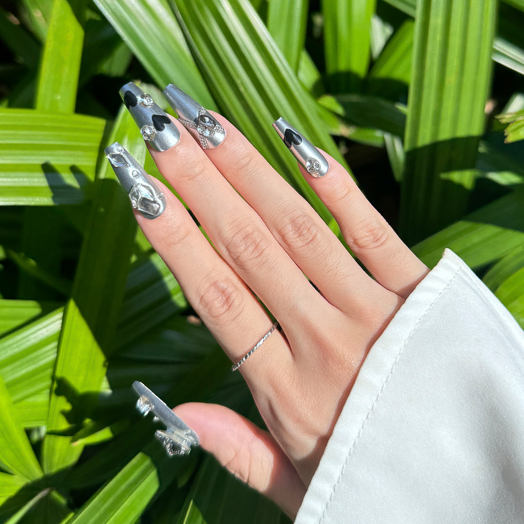 Heart Spell handmade long coffin-shaped metallic silver press-on nails with black heart design, gray heart gems, unique rhinestones, and metal chain accents
