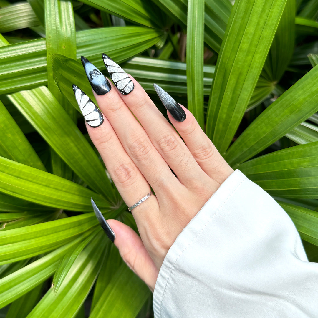 Stiletto-shaped press-on nails in black and silver, featuring glittery silver butterfly wing designs. Handmade with a dark, edgy, and unique butterfly-inspired style.