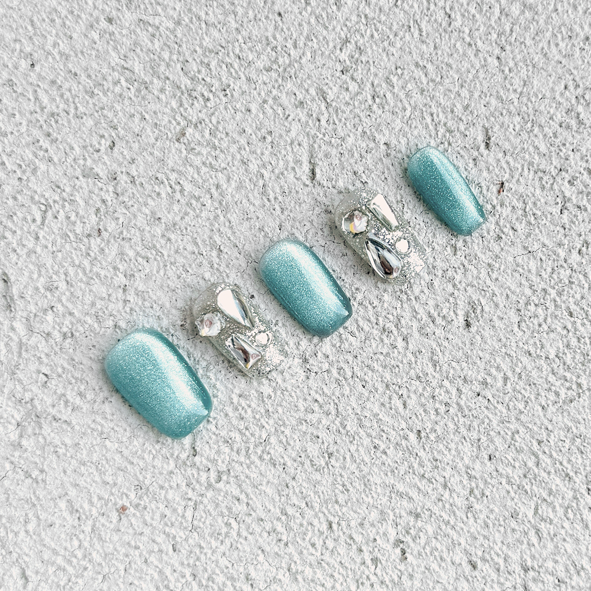 Handmade square-shaped press-on nails with blue-green cat-eye effect, silver accents, and unique crystal embellishments, inspired by vibrant ocean hues.
