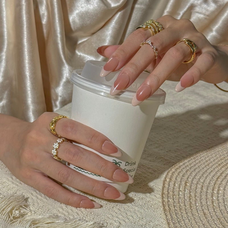 Fincloset almond-shaped press-on nails with French tips in subtle nude tones, paired with gold and floral rings. The nails are styled in a cozy, casual setting with a coffee cup, exuding an effortlessly polished and elegant aesthetic.