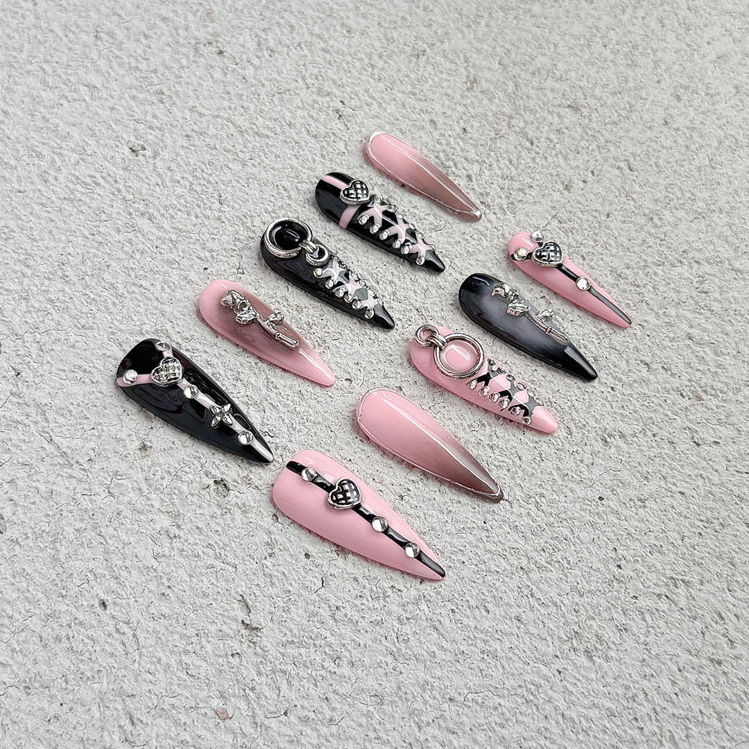 Handcrafted stiletto-shaped press-on nails in black and pink with metallic heart charms, silver rose details, and bold pink line patterns. Sweet, edgy, and daring.