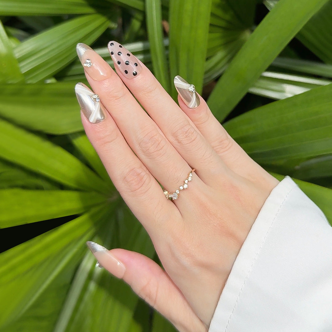 Almond-shaped press-on nails in nude and champagne gold with polka dots, gold brushed French tips, metallic mirror accents, white ribbon designs, and small gem embellishments. Luxurious and elegant.