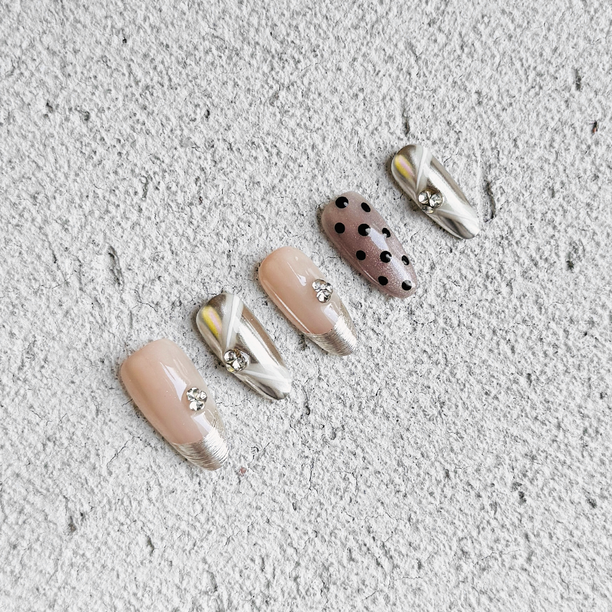 Almond-shaped press-on nails in nude and champagne gold with polka dots, gold brushed French tips, metallic mirror accents, white ribbon designs, and small gem embellishments. Luxurious and elegant.