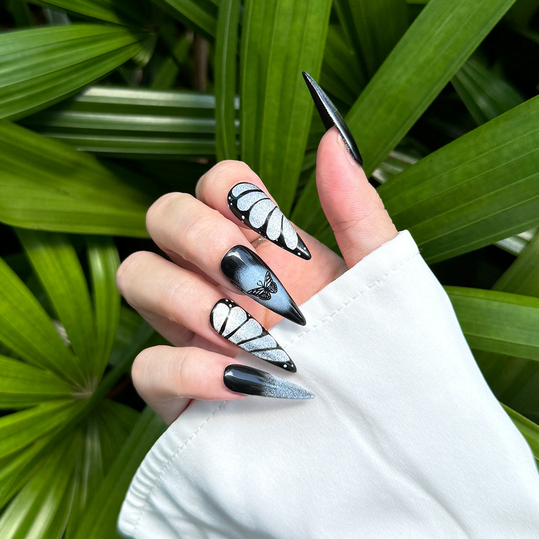 Stiletto-shaped press-on nails in black and silver, featuring glittery silver butterfly wing designs. Handmade with a dark, edgy, and unique butterfly-inspired style.