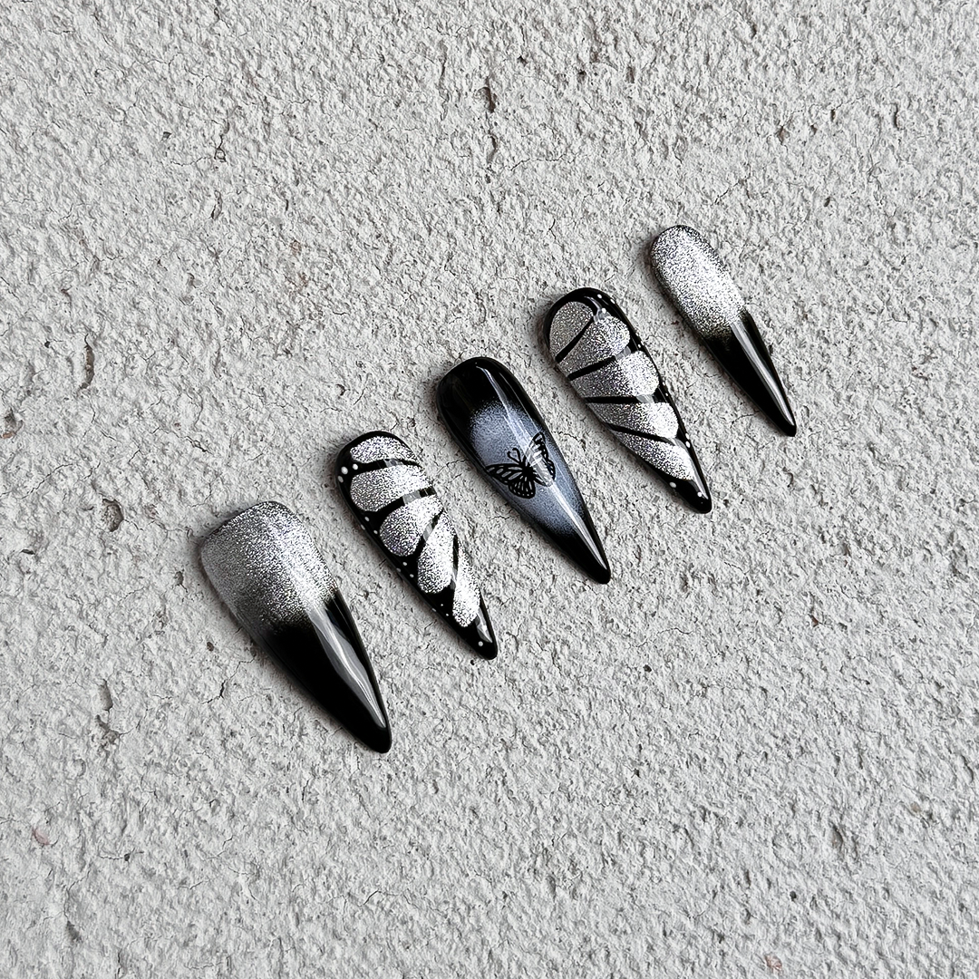 Stiletto-shaped press-on nails in black and silver, featuring glittery silver butterfly wing designs. Handmade with a dark, edgy, and unique butterfly-inspired style.