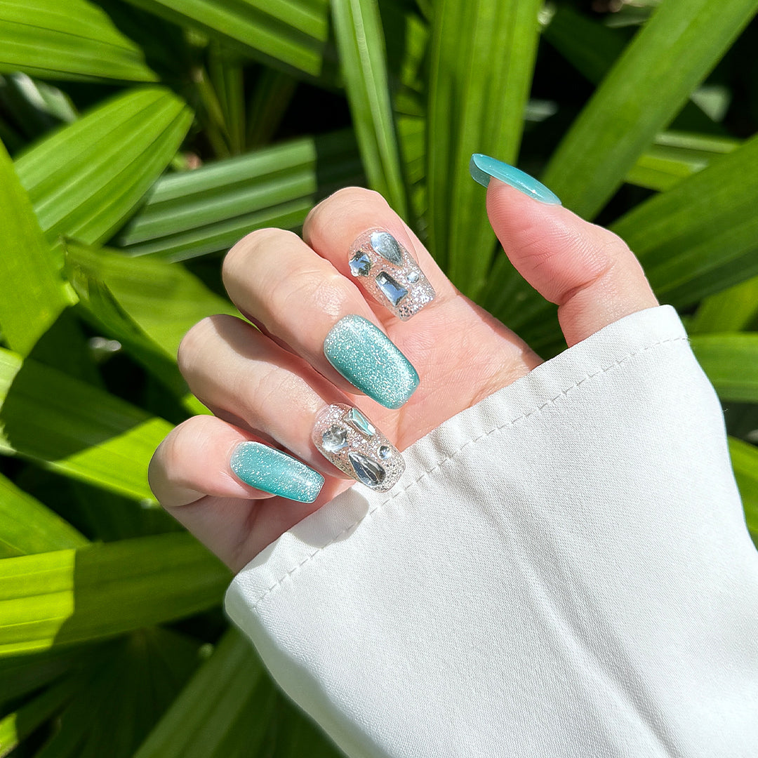 Handmade square-shaped press-on nails with blue-green cat-eye effect, silver accents, and unique crystal embellishments, inspired by vibrant ocean hues.