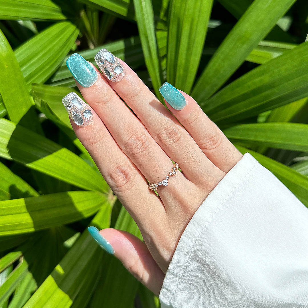 Handmade square-shaped press-on nails with blue-green cat-eye effect, silver accents, and unique crystal embellishments, inspired by vibrant ocean hues.