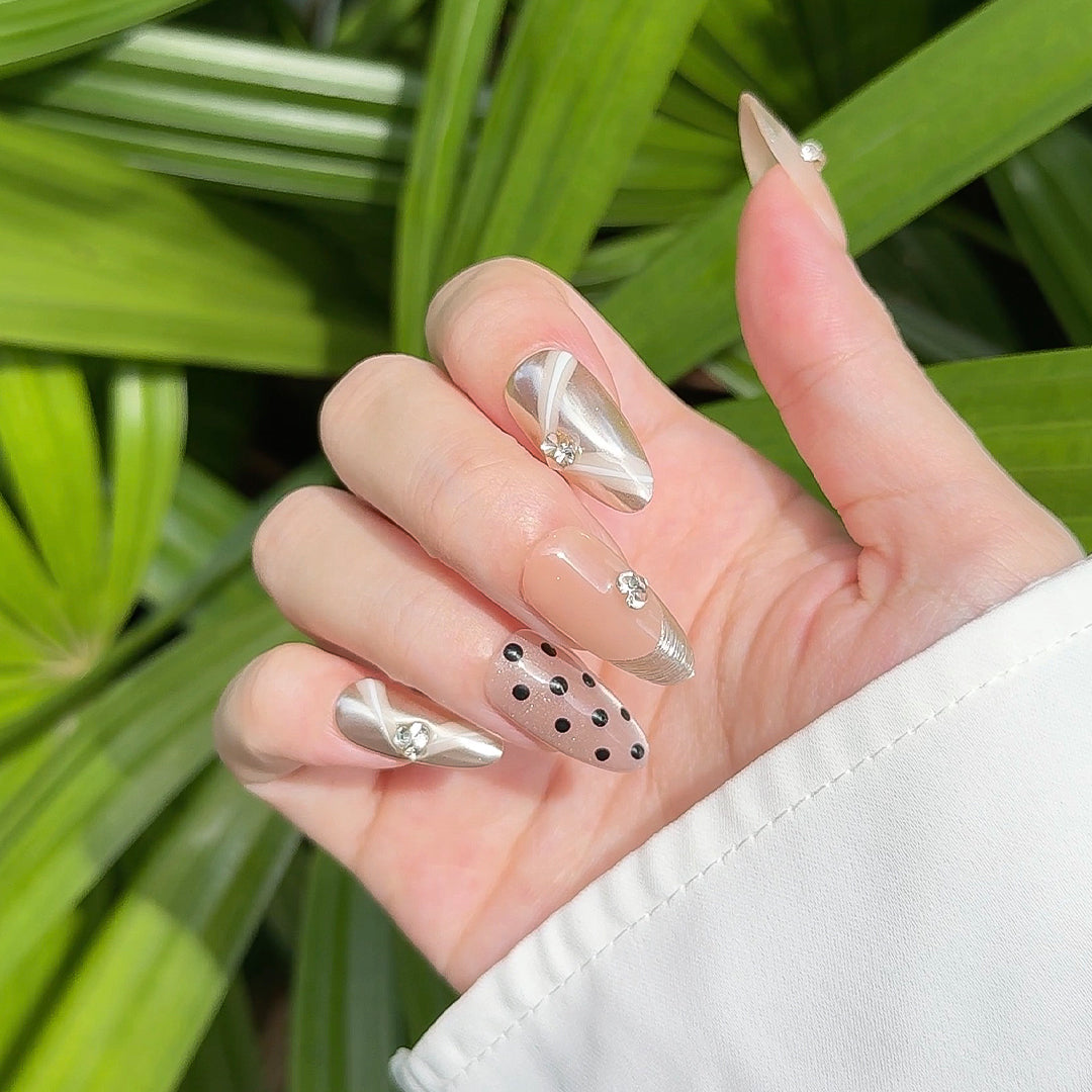 Almond-shaped press-on nails in nude and champagne gold with polka dots, gold brushed French tips, metallic mirror accents, white ribbon designs, and small gem embellishments. Luxurious and elegant.