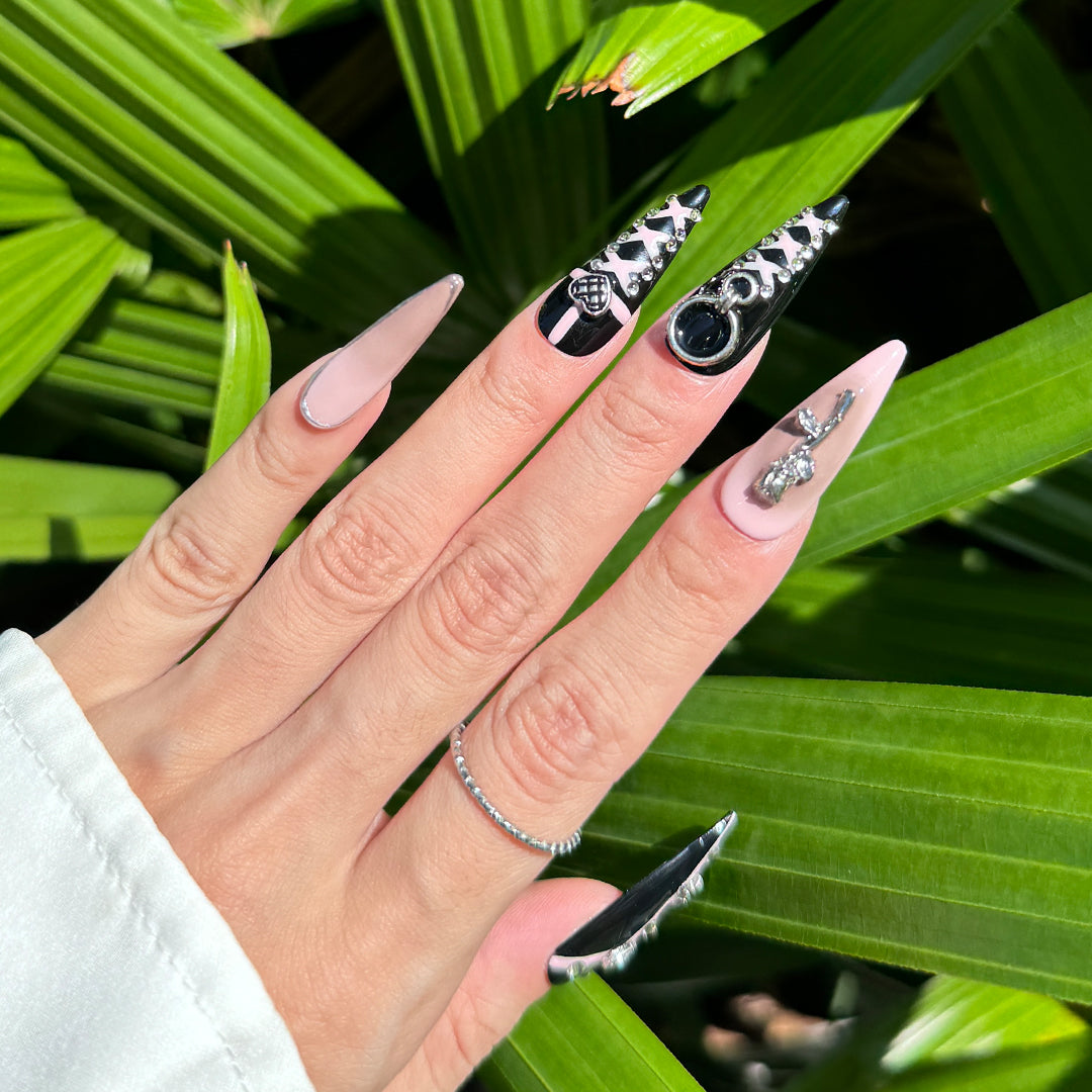 Handcrafted stiletto-shaped press-on nails in black and pink with metallic heart charms, silver rose details, and bold pink line patterns. Sweet, edgy, and daring.