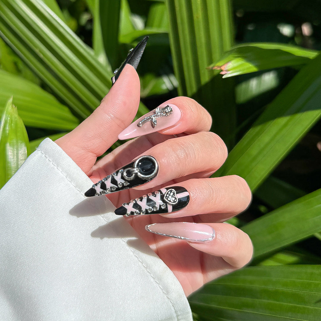 Handcrafted stiletto-shaped press-on nails in black and pink with metallic heart charms, silver rose details, and bold pink line patterns. Sweet, edgy, and daring.