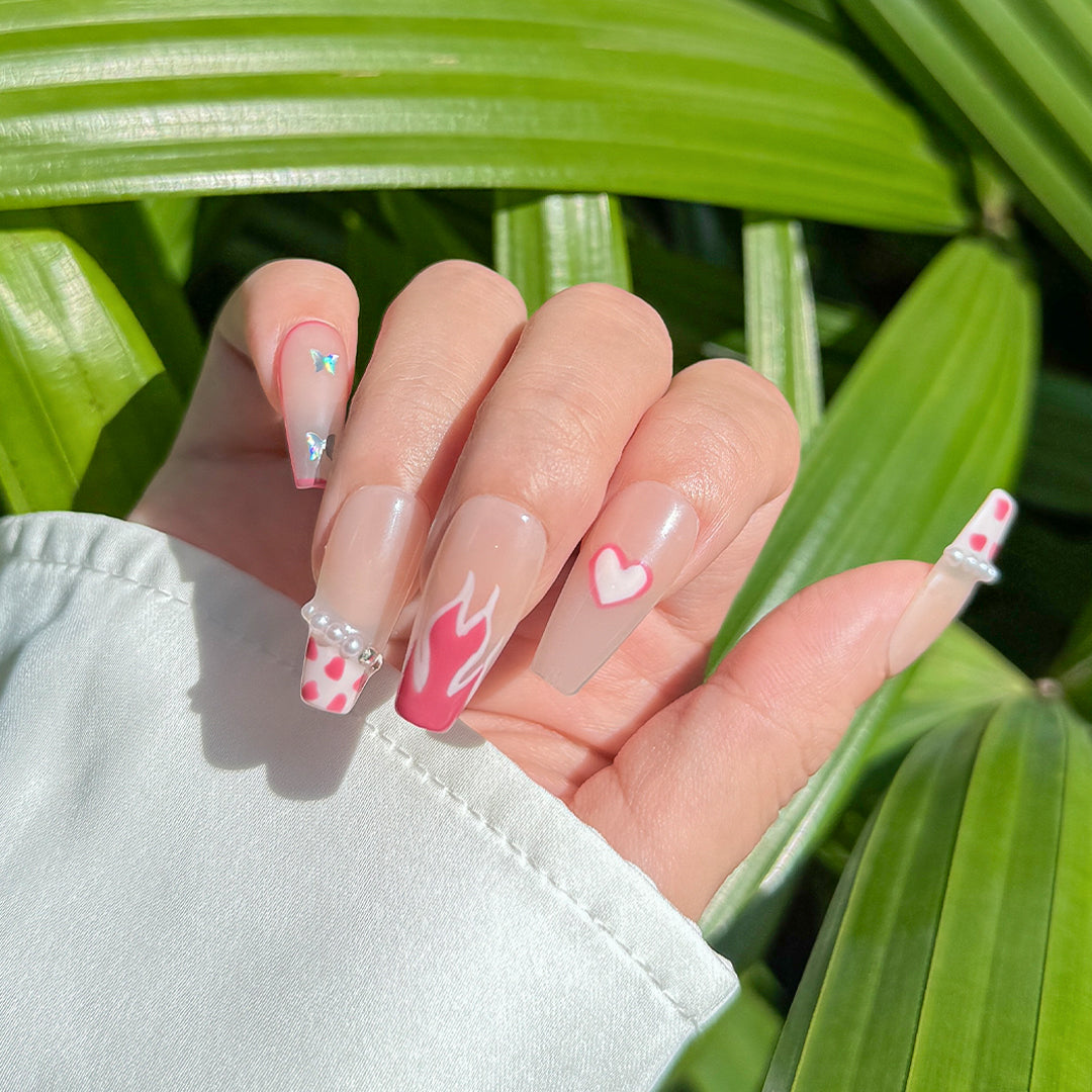 Handcrafted coffin-shaped press-on nails with translucent nude base, pink cow print, flame, heart designs, and pearl accents. Cute, playful, and elegant style.